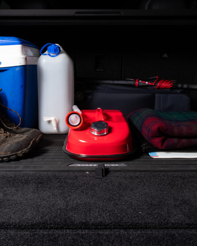Open car trunk with camping gear: blue cooler, white water jug with blue cap, red fuel can, folded umbrella, and plaid blanket inside.